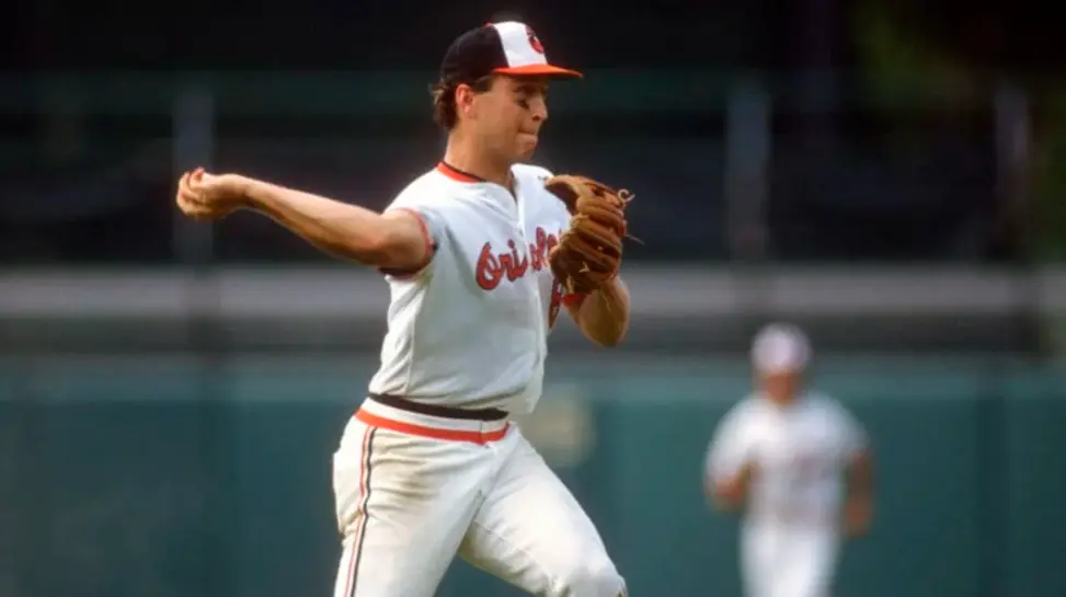 Baltimore Orioles player pitching during the 1966–1988 ‘Cartoon Bird’ era, wearing the white home uniform with orange and black trim and the iconic Cartoon Bird logo cap, representing the team’s championship dynasty period.