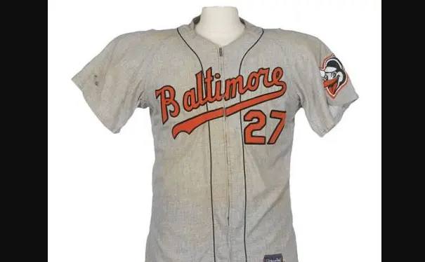 Baltimore Orioles road jersey from 1954–1965 in gray, featuring orange script ‘Baltimore’ with black trim and number 27 on the front, along with the early Oriole bird logo on the sleeve.