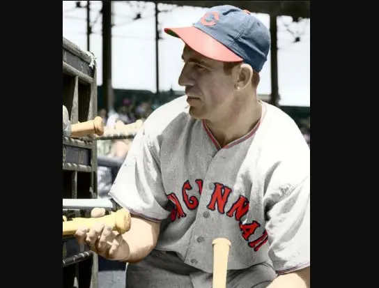 1910s–1950s Cincinnati Reds player wearing a gray flannel jersey with red ‘CINCINNATI’ lettering and a blue cap featuring the red wishbone ‘C’ logo, sitting in the dugout holding a bat