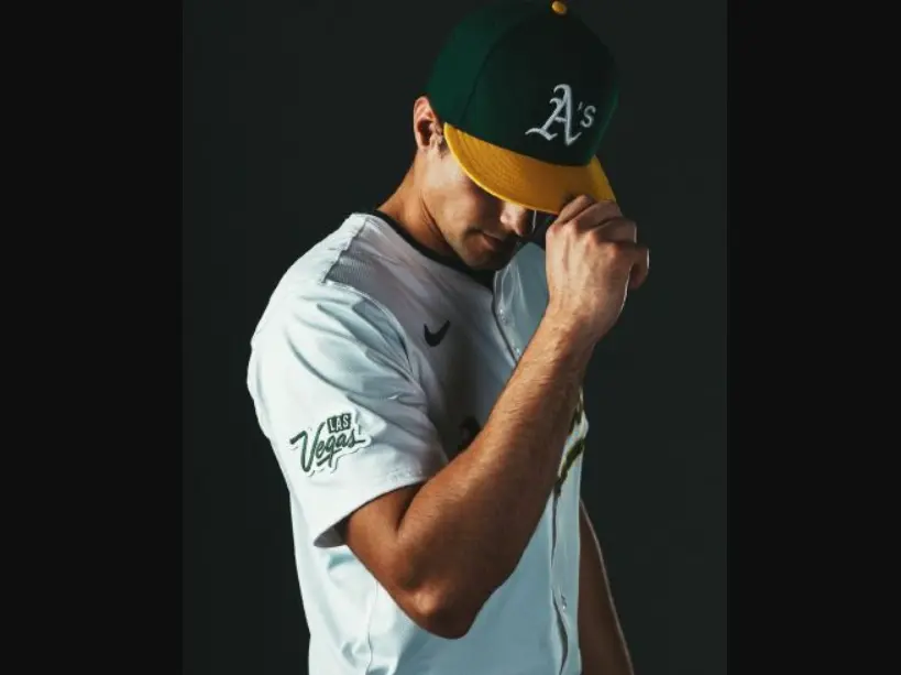 Oakland Athletics player wearing a modern white jersey with gold trim and a green cap featuring the ‘A’s’ logo, representing the 1993–present era with alternate and throwback designs