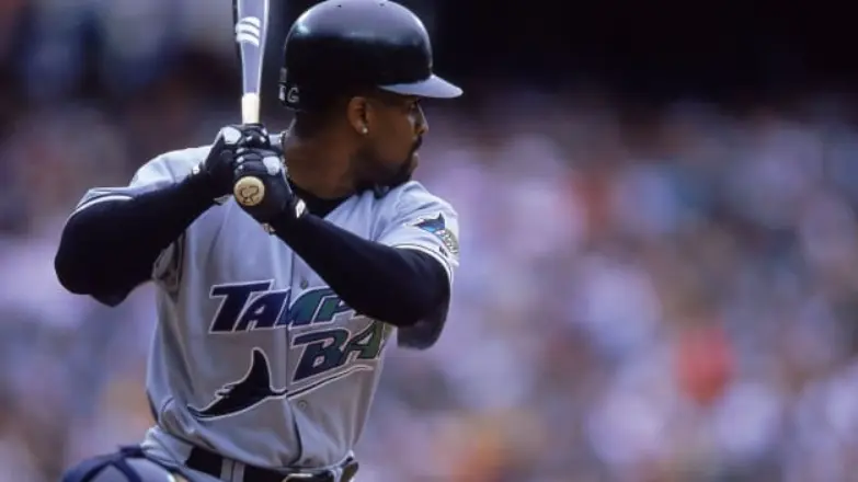 Tampa Bay Devil Rays player wearing the gray road uniform with a rainbow gradient ‘Devil Rays’ script across the chest and the colorful oval ray logo on the sleeve, representing the team’s bold inaugural look from the late 1990s
