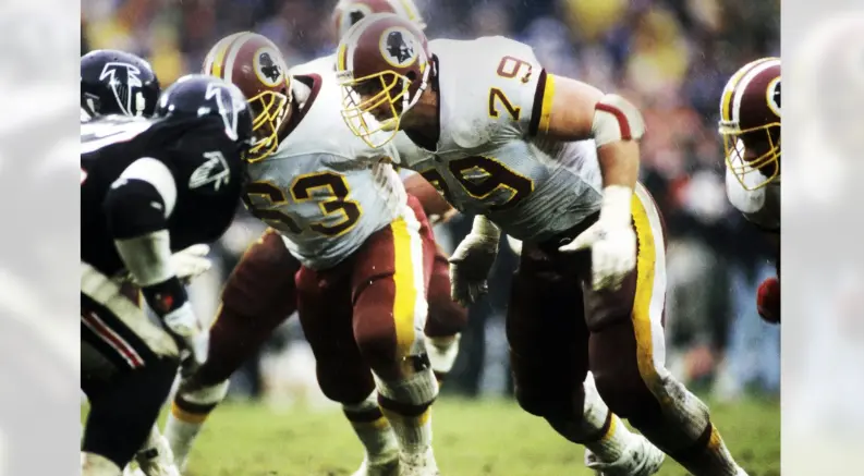 Action photo of Washington Redskins players from the 1981–2019 Joe Gibbs era wearing white jerseys with burgundy and gold accents and helmets featuring the classic circle logo with a Native American profile, symbolizing the team’s Super Bowl dynasty years