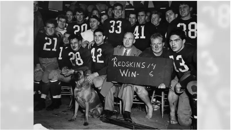 Black-and-white photo of the Boston Redskins team from 1933–1936 celebrating a win, with players wearing burgundy jerseys and a coach holding a sign reading 'Redskins 14 Win – 6,' representing the early Boston era before the franchise moved to Washington
