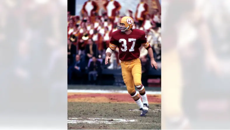 Color photo of a Washington Redskins player from the 1960s wearing a burgundy and gold uniform with the team’s first major helmet logo—a gold circle featuring a burgundy spear and feathers—symbolizing the era of stars like Sonny Jurgensen and Bobby Mitchell